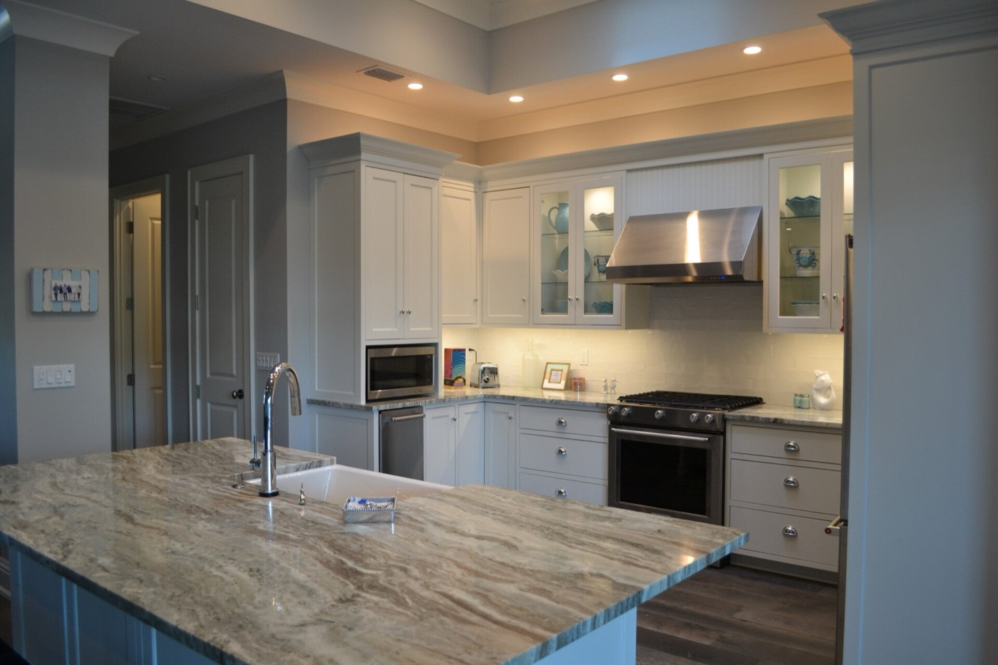 White kitchen with glass-front display cabinets, a stainless range hood, marble counters and under-cabinet lighting.
