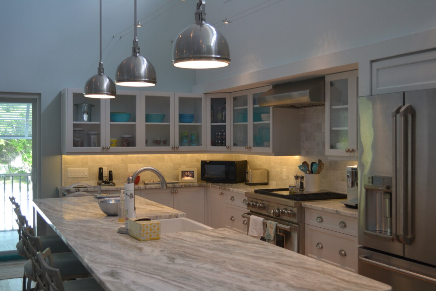 Kitchen with white cabinets, glass-front upper doors, a long marble island and polished nickel pendant lights.